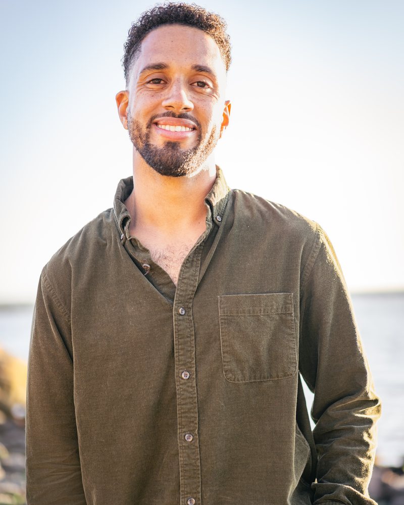 Nolan T. Krueger smiling, wearing an olive corduroy shirt, outdoors by the water.